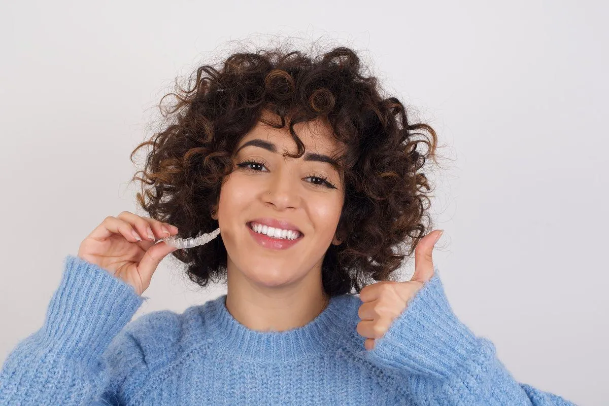 Young beautiful caucasian woman wearing blue knitted sweater standing against white studio background holding an invisible aligner braces and smiling. Dental healthcare concept